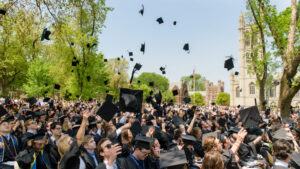 A large group of graduates in black robes and caps tossing their caps into the air