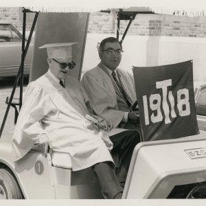 Two members of the Class of 1918 in graduation-style reunion gowns in golf cart at class reunion (Trinity College, Hartford Connecticut), 1968