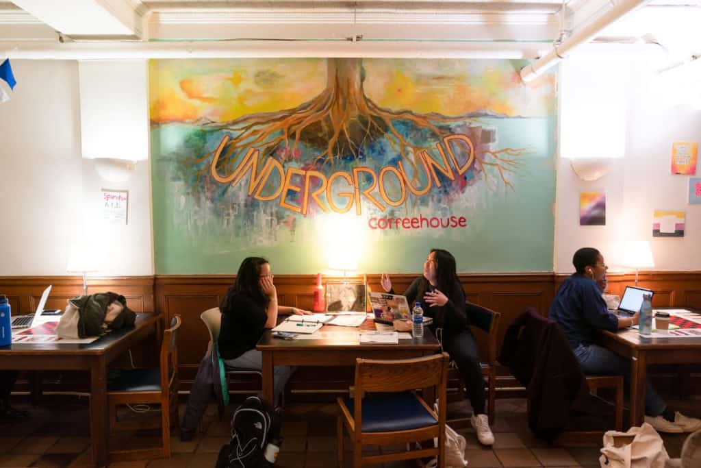 Two students work and socialize at a table with a large painting of the Underground Coffeehouse logo on the wall above them.