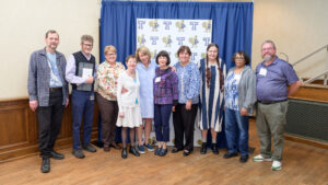 Group of retiring faculty and staff standing in front of a blue curtain and backdrop with Trinity logos